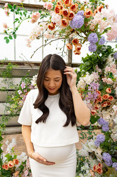 A pregnant woman rests one hand under her stomach and the other in her hair as she looks down at her stomach and is surrounded by flowers