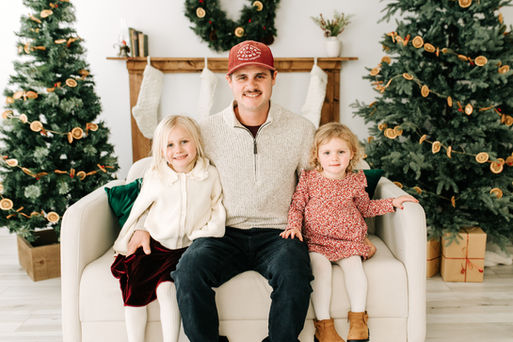 A man and two girls sit on a couch in between two Christmas trees