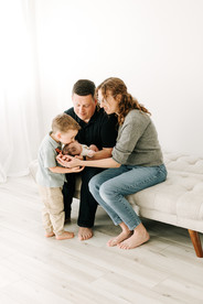 A man and woman sit on a couch and hold a newborn baby while a young boy stands in front of them and kisses the baby's head