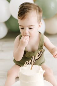 A one year old boy sits on the floor and touches a little white cake as green and white balloon arch is behind him