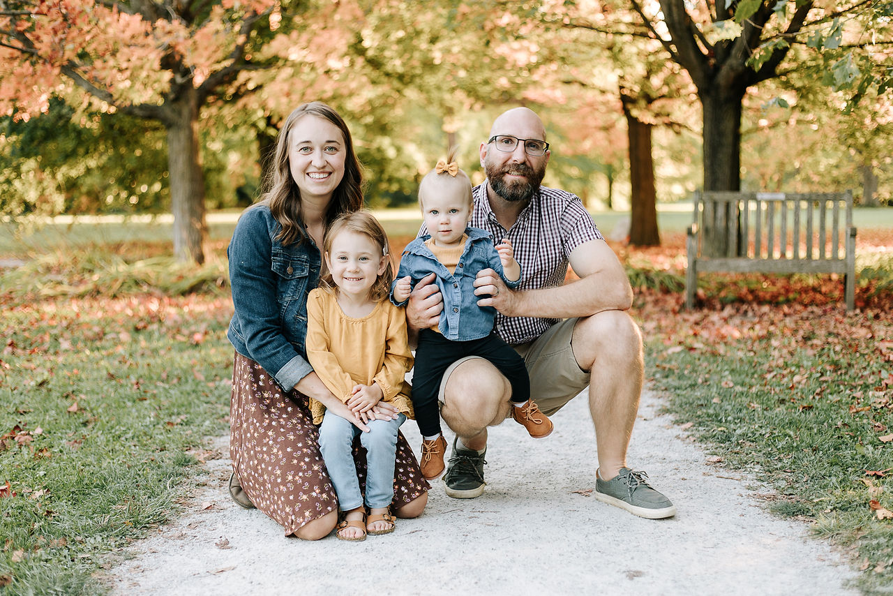 A woman and man crouch down and hug a young girl and toddler girl under autumn trees