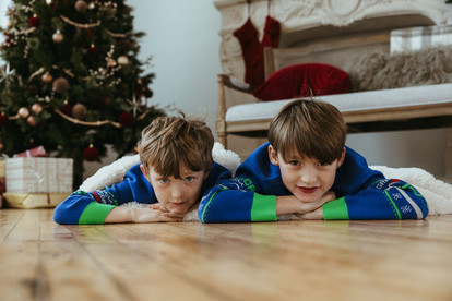 Two boys lie on their stomachs with their chins resting on their hands in front of a Christmas tree