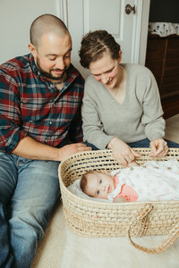 A man and woman sit on the floor and look down at a baby lying in a bassinet