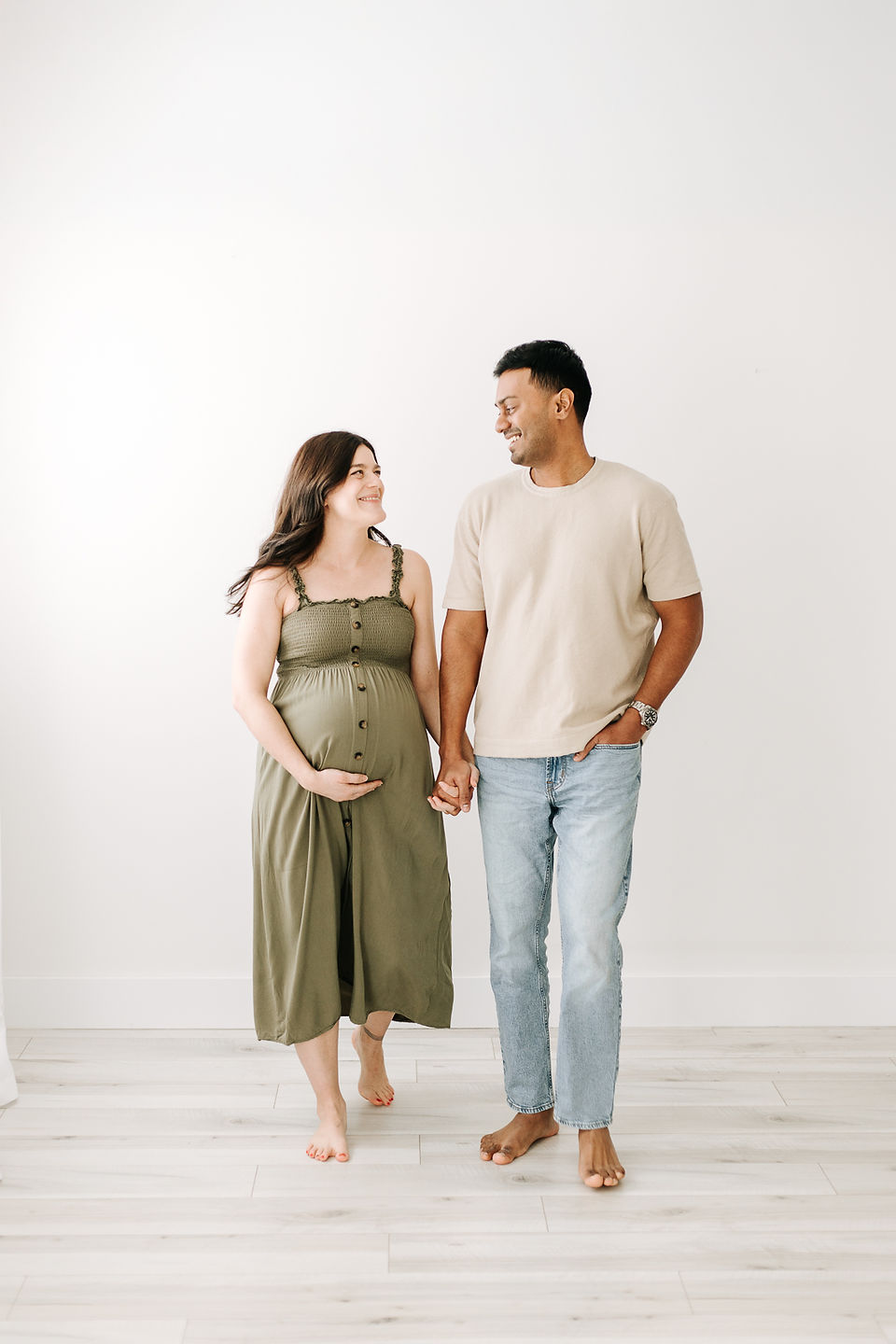 a woman in a green dress cradles pregnant stomach and walks as she holds hands with a man in jeans and a t shirt looking at each other
