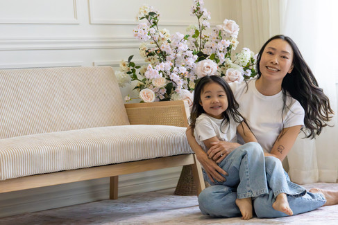 A woman sits on the floor and a young girl sits in her lap and behind them is a couch and floral arrangement
