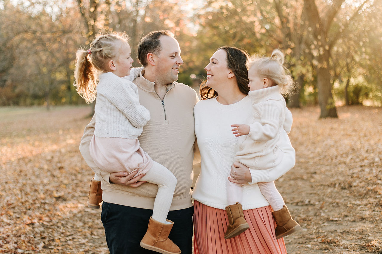 A man and woman each hold a little girl in their arms as they smile on a path under trees in the fall