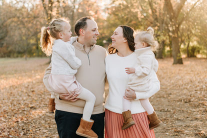 A man and woman each hold a little girl in their arms as they smile on a path under trees in the fall