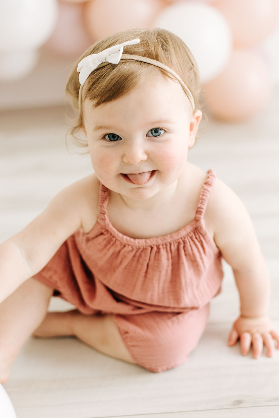 A one year old girl in a pink dress and white hair bow sits on the floor in front of a balloon garland and sticks out her tongue