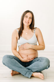 pregnant woman sits on the floor in jeans and a bra holding her stomach and looking straight ahead