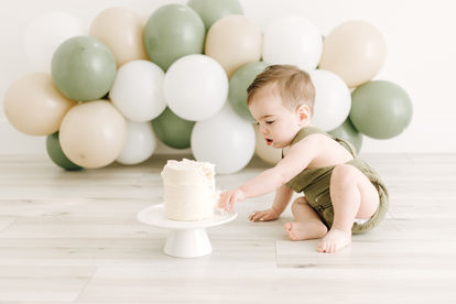 A one year old boy sits on the floor and touches a little white cake as green and white balloon arch is behind him