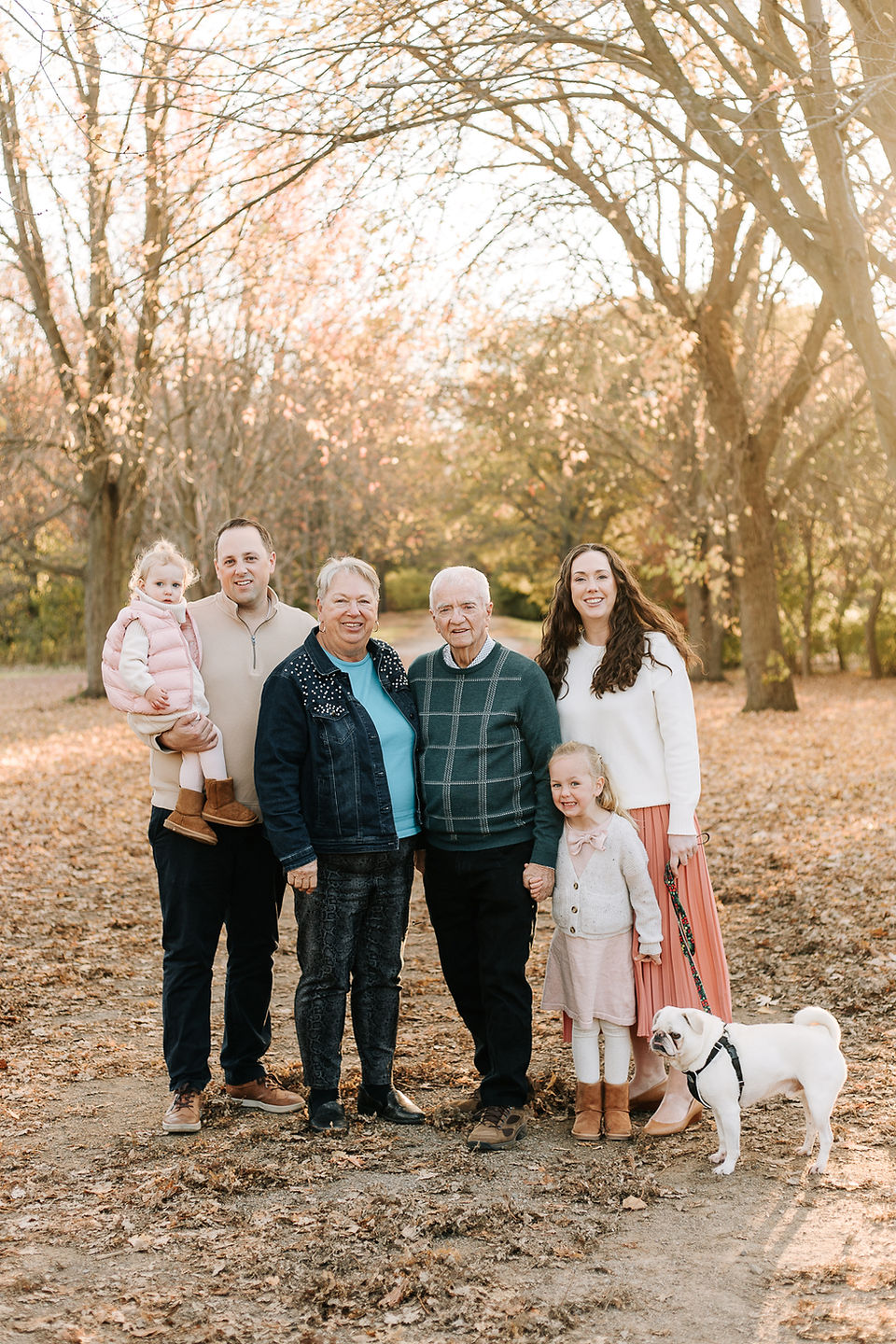 A couple stand and smile in a forest with two girls, a dog, and an older couple