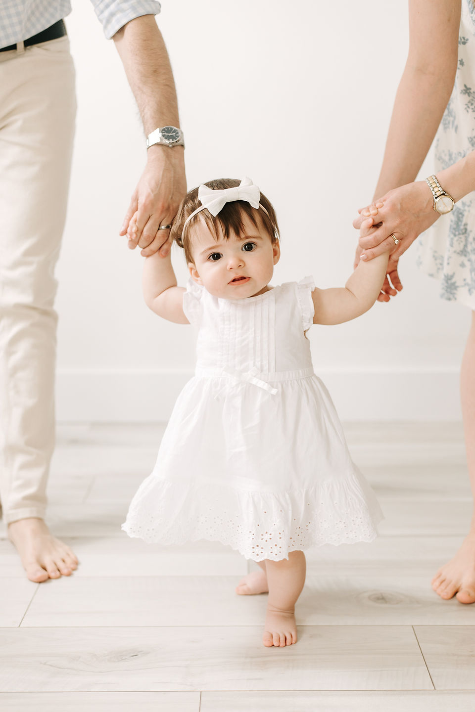 One year old girl in a white dress and hair bow holds hands of a man and woman