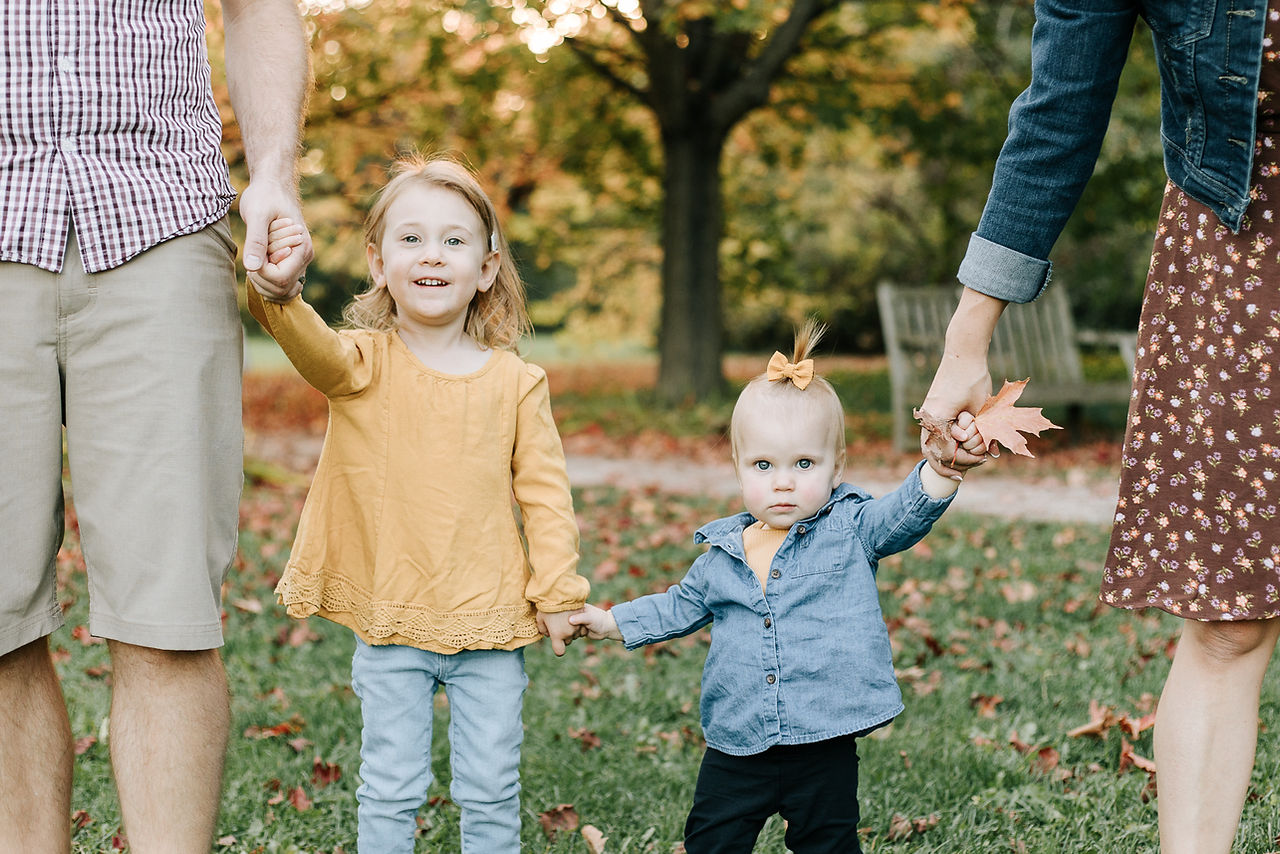 A woman and man hold hands with a young girl and toddler girl under autumn trees