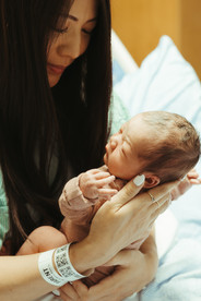 A woman in a hospital gown sits on a hospital bed and holds a newborn baby in her arms