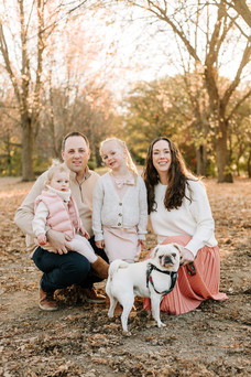 A woman and man crouch down as two young girls and a dog stand by them under trees in fall