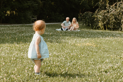 A one year old girl stands and looks at an older couple who are sitting on a blanket in grass