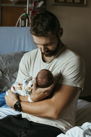 A man sits on the edge of a hospital bed cradling a newborn baby and looks down at the baby