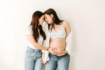 two woman stand next to each holding hands and resting their foreheads together, one of the woman is in a white t shirt and jeans and the second woman is in jeans a calvin klein bra and white button down shirt