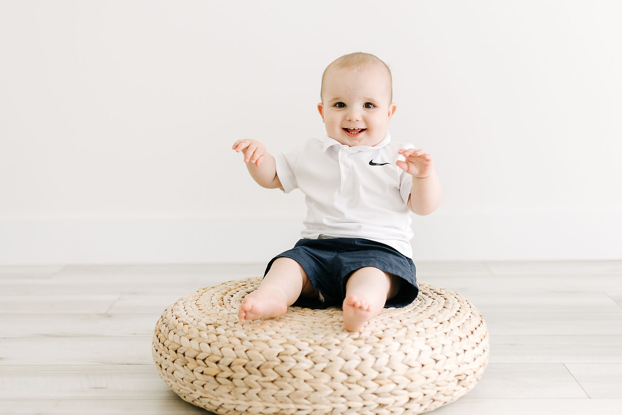 A one year old boy smiles as he sits on a rattan pouf