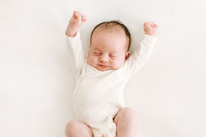 A newborn baby in a long sleeve white onesie stretches her arms