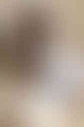 A pregnant woman sits on the edge of a bed and looks down at a onesie and baby booties and rests one hand on her pregnant stomach