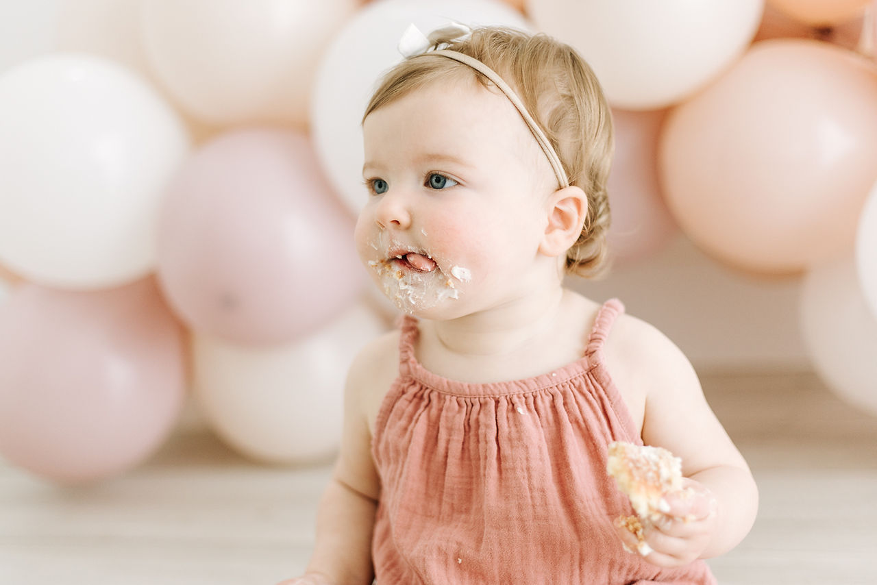 A one year old in a pink dress and hair bow licks cake off of her face as she sits in front of a balloon garland