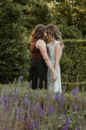 Two woman hold hands and rest their foreheads together as they stand in between a tall hedge and some lavendar