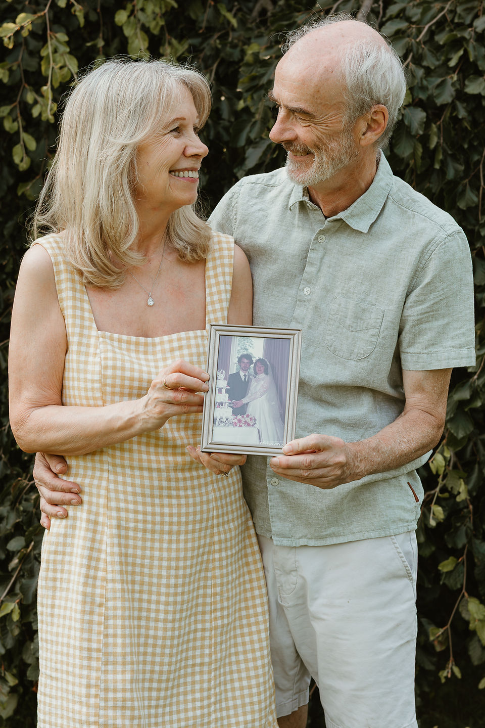 An older woman and man look at each other and smile as they hold a photograph of their wedding day