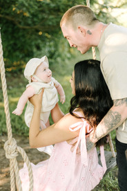 A woman sits on a swing and holds a baby in her arms while a man stands behind her with his hands on her back and looks down at the baby
