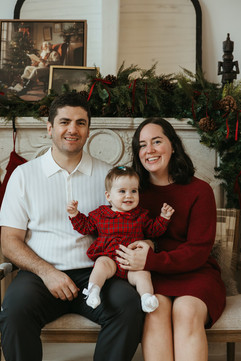 A man and woman with a baby girl on their lap and sit on a couch in front of a fireplace with a Christmas garland and photos on it
