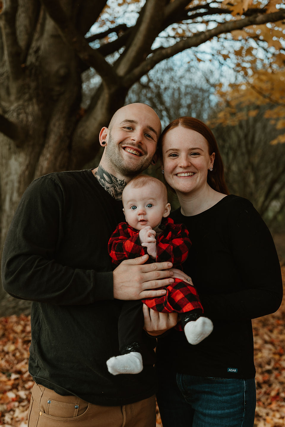 A man and woman stand together as they hold a baby under an autumn tree