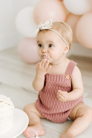 A one year old sits on the floor in front of a balloon arch and eats a small white cake