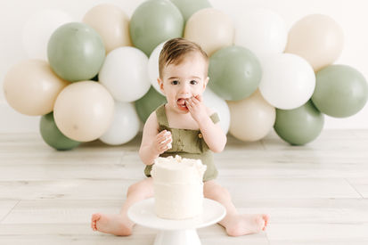 A one year old boy sits on the floor and touches a little white cake as green and white balloon arch is behind him
