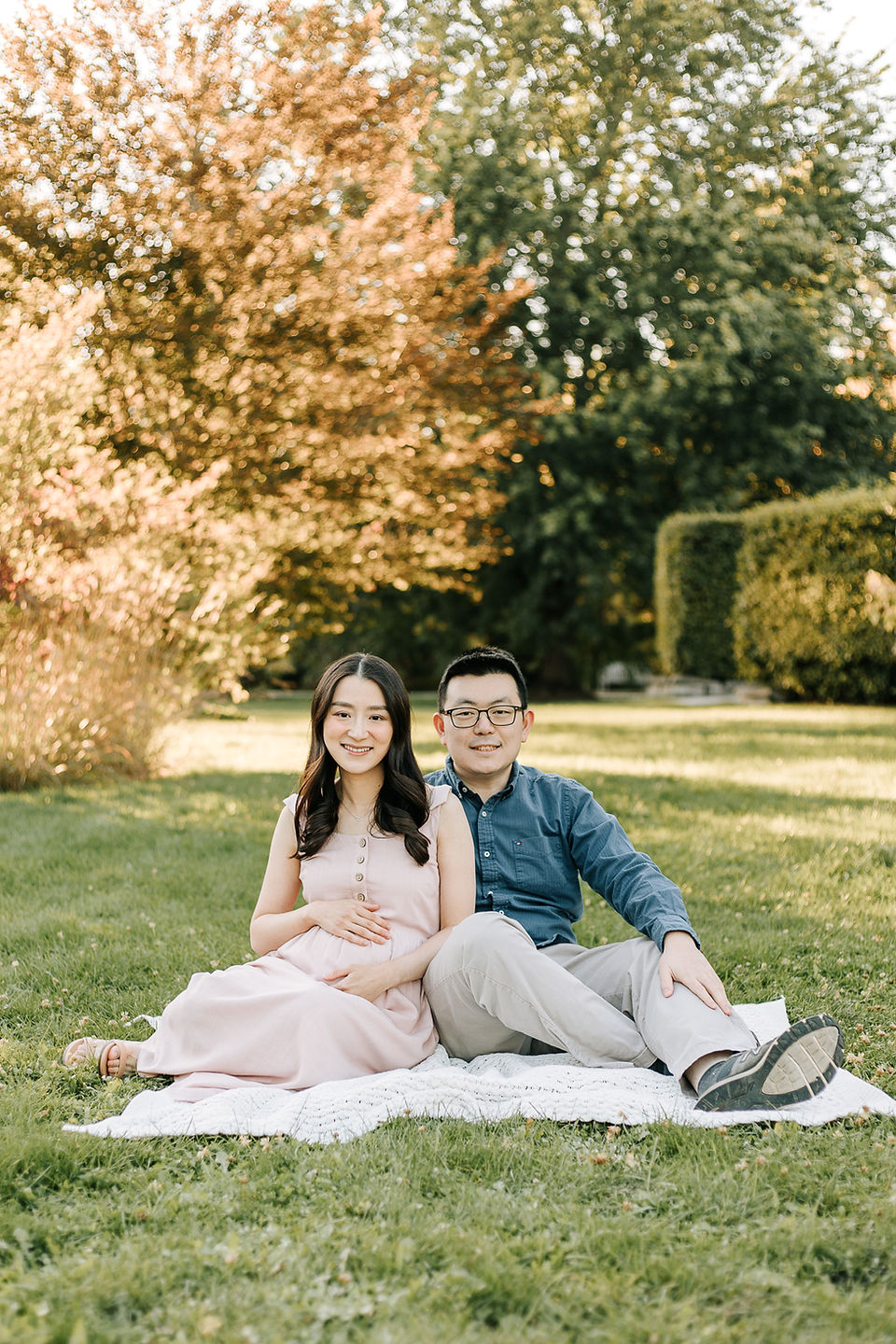 A pregnant woman and man sit on a blanket on grass in a park