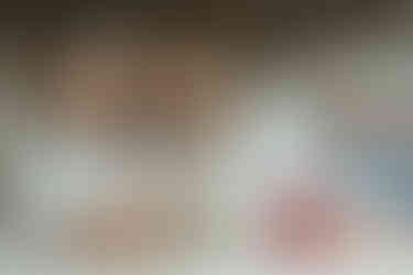 A baby lies on a bed asleep as her parents lie behind her and look down at her