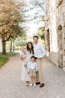 A man and woman stand with their arms around each other as the woman holds a young boy in her arms and another boy stands in front of them