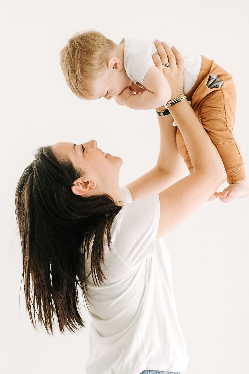 A woman in a white t shirt holds a one year old boy in the air and smiles at him