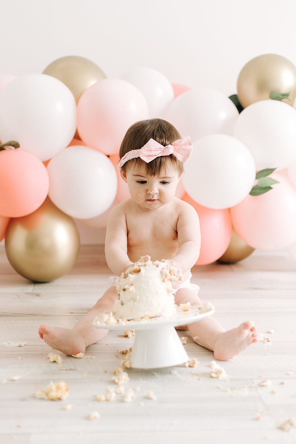 A one year old girl in a pink hair bow and diaper sits behind a small white cake with her hands in the cake
