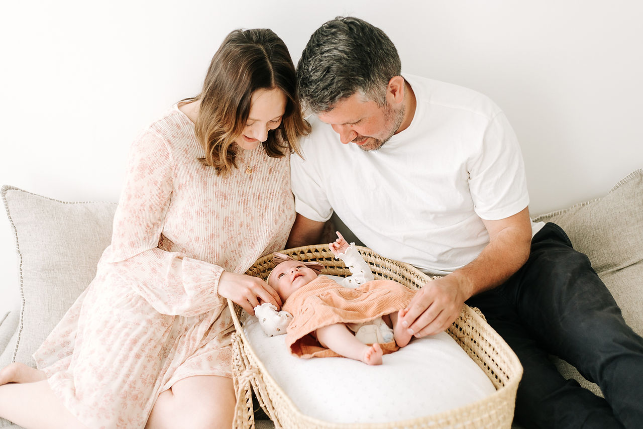 woman and man sit beside each other on a couch and look down at the baby in a bassinet