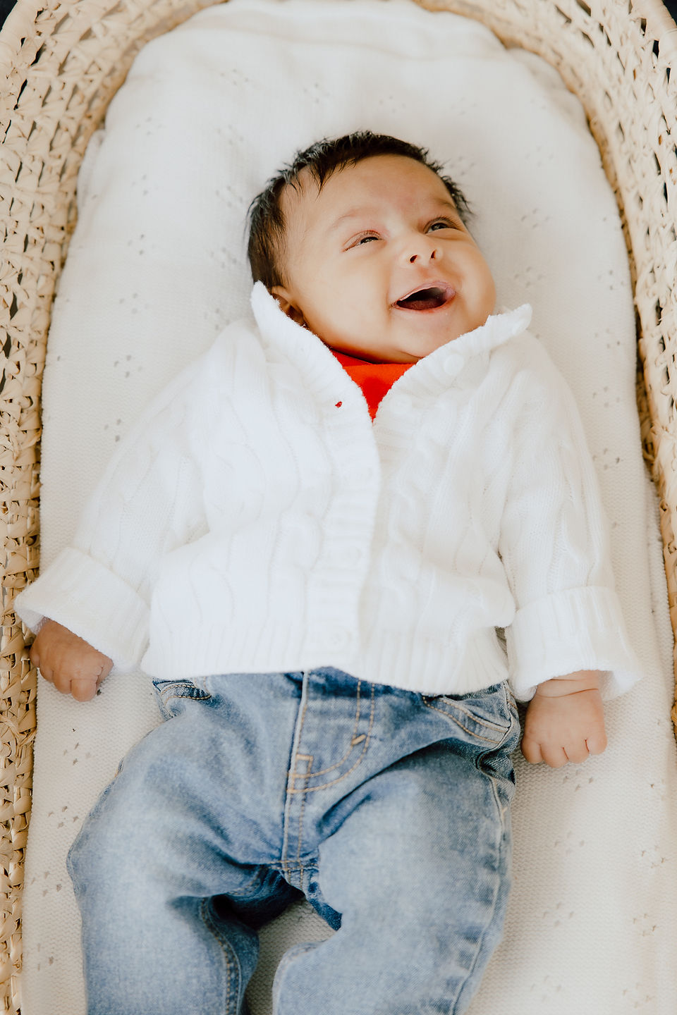 A smiling baby lies in a bassinet in jeans, a red tshirt and white sweater