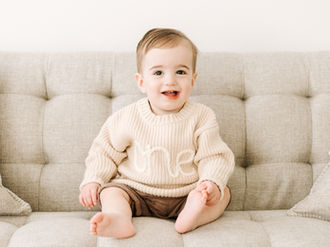A one year old sits on a couch and is wearing a beige sweater that says ONE