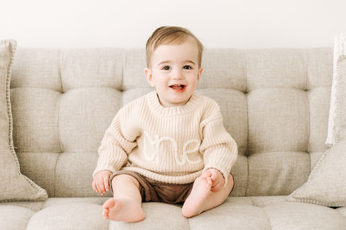 A boy in a beige sweater that spells one sits on a couch and smiles