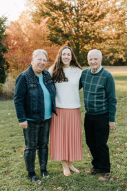 A woman stands between an older man and woman with her arms around them as they all smile