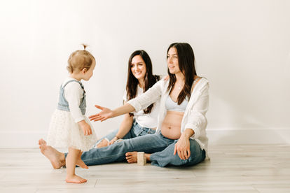 A toddler girl faces two smiling woman sitting on the floor, one of whom is reaching her arm out towards the toddler