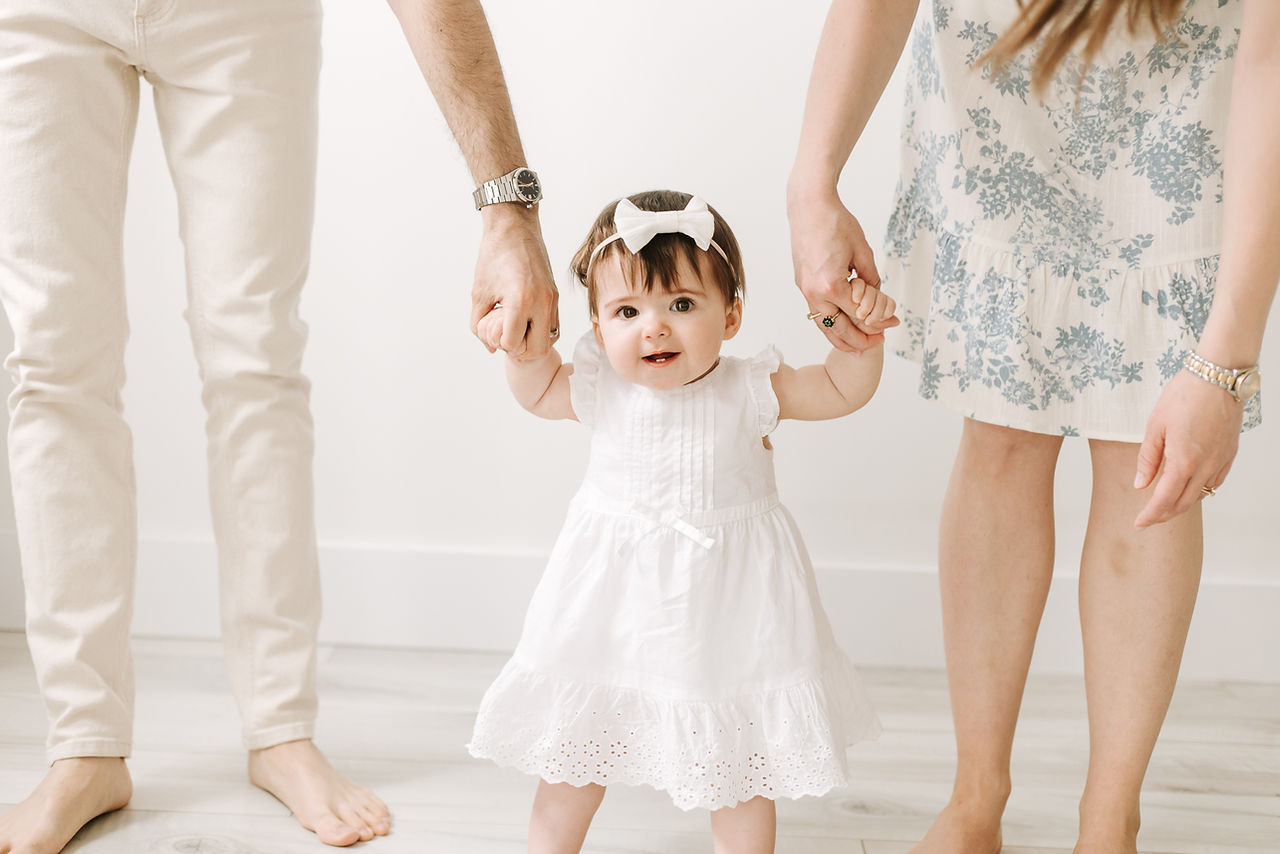 One year old girl in a white dress and hair bow holds hands of a man and woman