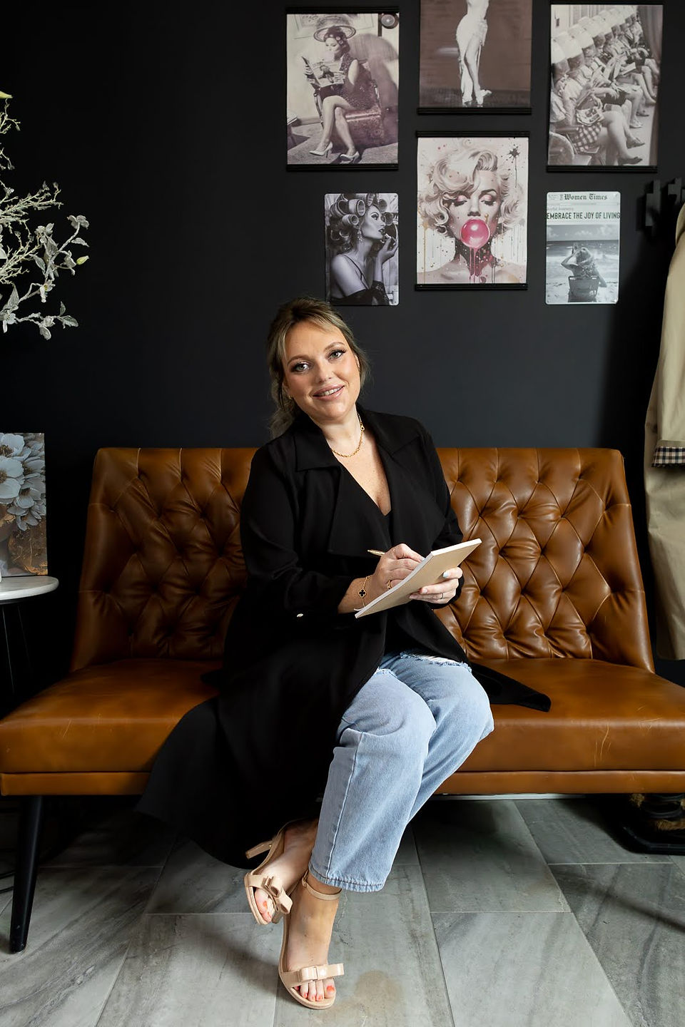 A woman in a black jacket and jeans sits on a brown couch as she holds a pen and notebook