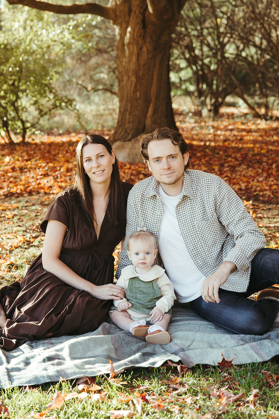 A man, woman, and baby sit on a blanket in a park in autummn