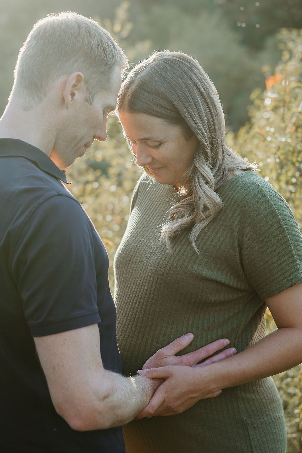 A pregnant woman and a man rest their hands on her stomach as they stands in a field