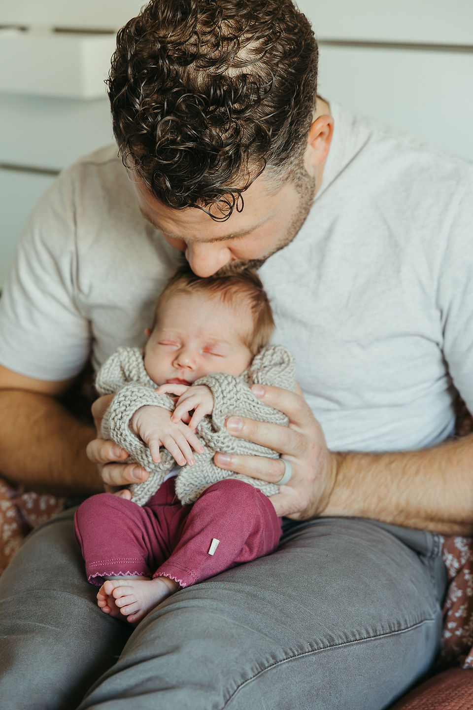 A man sits on a bed and kisses the top of a baby's head that is sitting on his lap