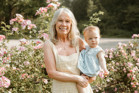 An older woman holds a one year old girl and smiles as she stands in front of flowers